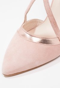 Rosa mockasin högklackad sko med spetsig tå och en metallisk rosaguld accent längs framkanten. Har korskorsande vristremmar.