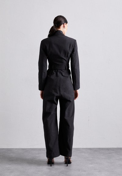 Salopetă neagră, croită, cu talie ajustată, mâneci lungi și pantaloni evazați. Prezintă o textură netedă și un design bine structurat.