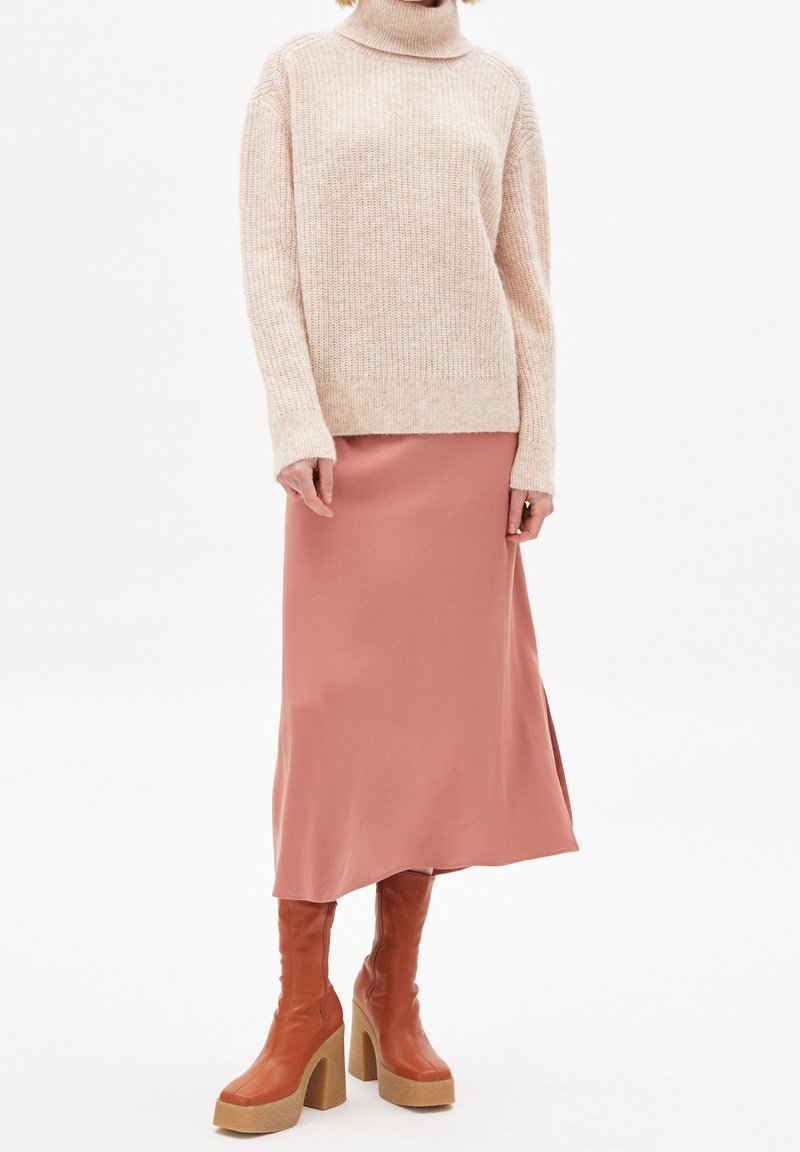 Pull à col roulé en tricot beige, jupe midi en satin rose, et bottes en cuir marron au genou avec des talons épais, créant une tenue superposée.