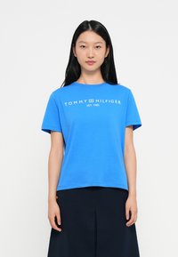 Kobieta z długimi czarnymi włosami, ubrana w niebieską koszulkę Tommy Hilfiger i czarną spódnicę, stojąca na tle gładkiego białego tła.
