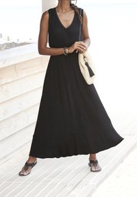 Svart ärmlös maxiklänning med V-ringning, plisserad design, tillsammans med en vävd beige väska med en svart tofsdetalj och platta sandaler.