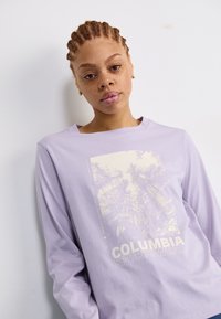 Šviesiai violetinė ilgomis rankovėmis marškinėliai su grafiniu spaudiniu, kuriame pavaizduoti medžiai ir užrašas "COLUMBIA SPORTSWEAR", pagaminti iš minkštos medžiagos. Atsipalaidavęs siluetas.