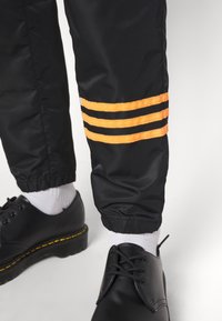 Black nylon pants with elastic cuffs featuring three orange and black striped accents. Pair of black lace-up shoes with white socks.