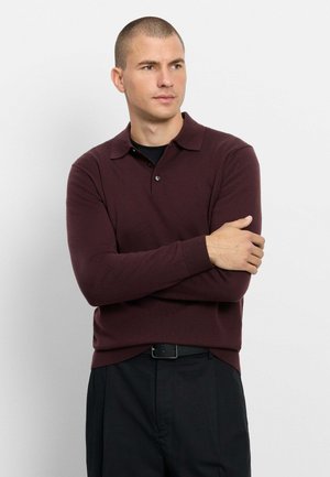 Mann mit kurzen Haaren, der ein dunkelburgunderfarbenes Langarm-Poloshirt und schwarze Hose trägt, verschränkt die Arme und schaut zur Seite.