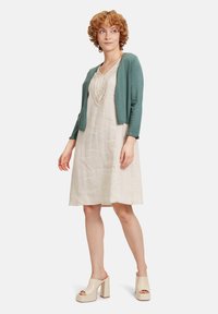 Cardigan verde corto sopra un vestito di lino beige con scollatura ricamata, abbinato a mule beige con tacco a blocco.