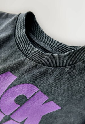 Sweatshirt gris à col rond, avec un col côtelé, présentant un grand design de texte graphique violet sur le devant. Texture de tissu douce.