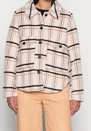 Vrouw draagt een beige en zwart geruit jasje met knopen, twee klepzakken aan de voorkant en een beige broek.