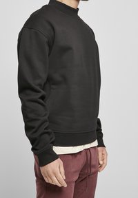 Sudadera negra con cuello alto, mangas largas y puños acanalados. Textura suave; usada sobre una camisa crema y combinada con pantalones color marrón rojizo.