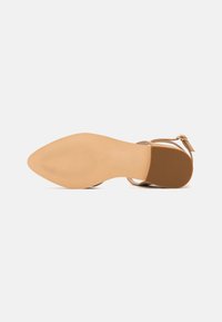 Sandal de punta beige con suela plana, tirante en el tobillo y pequeña hebilla, mostrado desde el lado de la suela sobre un fondo blanco liso.
