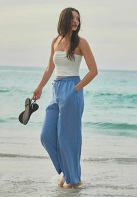 Pantaloni vaporoși de culoare albastru deschis, cu talie ajustabilă, asortați cu un top alb fără bretele. Persoană ținând în mână șlapi negri pe plajă.