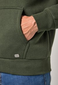 Grüne Sweatshirt mit strukturiertem Stoff, Kängurutasche, gerippten Bündchen und einem kleinen Metalllogo-Akzent nahe dem Saum.