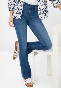 Blauwe spijkerbroek met uitlopende pijpen en een hoge taille, voorzien van een vijf-pocket design, gecombineerd met witte sandaaltjes met bandjes en een gemusterde top.