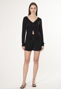 Cardigan nero con scollatura a V, maniche lunghe e bottoni frontali, abbinato a pantaloni corti neri. Il tessuto appare leggero e morbido.