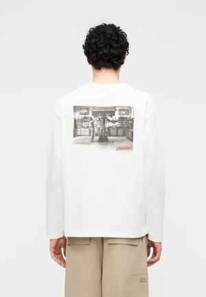 Άτομο με σγουρά μαλλιά που φοράει άσπρο πουκάμισο μακρυμάνικο με ασπρόμαυρη σκηνή συνεργείου γκαράζ και μπεζ παντελόνι με ένθετο λογότυπο Dickies.