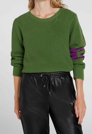 Femme portant un pull en maille vert avec une manche retroussée révélant une couche intérieure violette, associée à un pantalon en cuir noir à cordon de serrage.