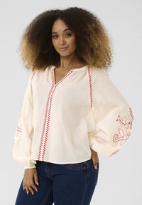 Witte blouse met rode geborduurde accenten en stiksels. Beschikt over een V-hals, luchtige mouwen en bloemenpatronen op de mouwen.