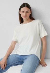Witte t-shirt met korte mouwen, relaxte pasvorm en opgerolde mouwen, gecombineerd met lichtblauwe jeans, tegen een neutrale achtergrond.