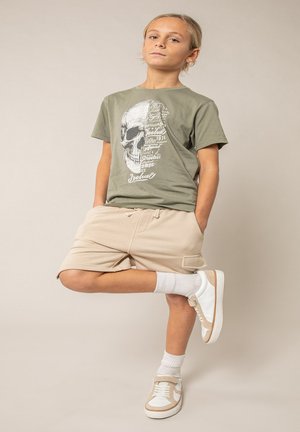 Enfant debout avec une jambe levée, portant un t-shirt vert olive avec un graphique de crâne, un short beige, des chaussettes blanches et des baskets blanches.