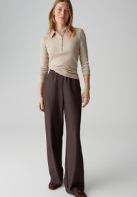 Vrouw met blond haar die een beige geribd poloshirt met lange mouwen en donkerbruine, wijd uitlopende broek draagt, staand tegen een effen achtergrond.