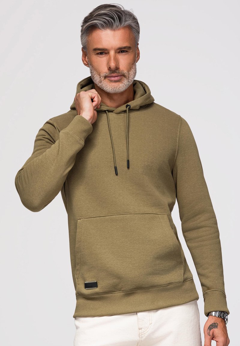 Mann mit grauen Haaren und Bart, der einen olivgrünen Hoodie und weiße Hose trägt, hält den Hoodie-Kragen mit der linken Hand.