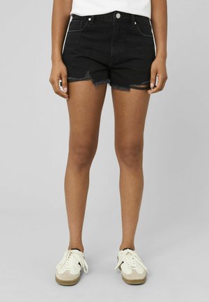 Personne portant un short en jean noir déchiré, une chemise blanche et des baskets blanches, sur un fond gris uni.