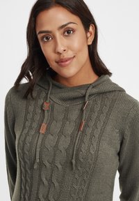 Sweat à capuche en maille cable en vert olive, arborant des motifs texturés, des cordons avec des accents en cuir, et une coupe décontractée et confortable.