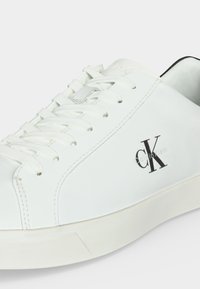 Λευκό sneaker χαμηλού τύπου με λευκά κορδόνια, μαύρο λογότυπο "CK" και κείμενο "Calvin Klein" στο πλάι, με λευκή λαστιχένια σόλα.