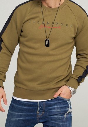 Man in een olijfgroen sweatshirt met zwarte strepen, tekst "Jack & Jones Originals", blauwe distressed jeans, een zilveren horloge en een zwarte hanger ketting.
