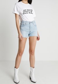 Camiseta blanca de algodón con texto negro, combinada con pantalones cortos de mezclilla azul claro. El conjunto se complementa con botas vaqueras blancas.