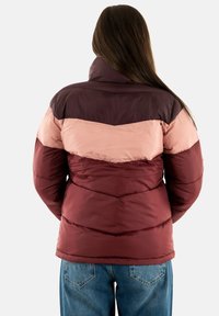 Giacca a vento con colletto alto, presenta un design colorato a blocchi in bordeaux scuro, rosa chiaro e marrone, con una texture trapuntata e un orlo arrotondato.