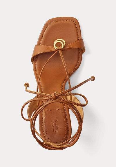 Καφέ δερμάτινο σανδάλι με επίπεδη σόλα, τετράγωνη μύτη, χρυσή στρογγυλή αγκράφα και μακριά λουριά που τυλίγονται γύρω από τον αστράγαλο και δένονται σε φιόγκο.