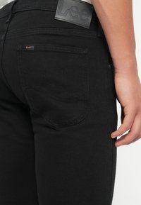 Fekete farmer nadrág, slim fit szabással, bőrszíjjal a deréknál és diszkrét varrási részletekkel ellátott hátsó zsebbel.