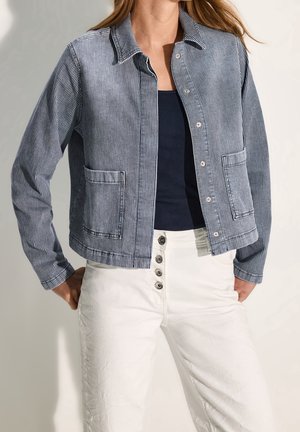Femme portant une veste boutonnée rayée bleue et blanche sur un haut noir, associée à un pantalon blanc taille haute avec quatre boutons visibles.