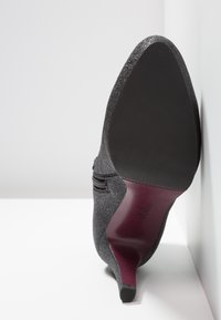 Chaussure à talon haut à bout pointu, grise pailletée, reposant sur le côté, montrant la semelle noire et le talon élancé bordeaux avec le logo de la marque embossé.