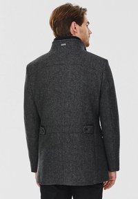 Szary wełniany płaszcz o wyrafinowanej konstrukcji, z wysokim kołnierzem, dopasowanym krojem i guzikami na plecach, wykonany z teksturowanej tkaniny i charakteryzujący się czystymi liniami.