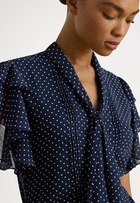 Nő tengerészkék pöttyös blúzban, fodros ujjakkal és nyakkendővel, lefelé néz egy egyszínű világos háttér előtt.