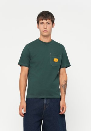 Green cotton t-shirt with a round neckline, short sleeves, and a chest pocket featuring a yellow label. Worn with dark blue jeans.