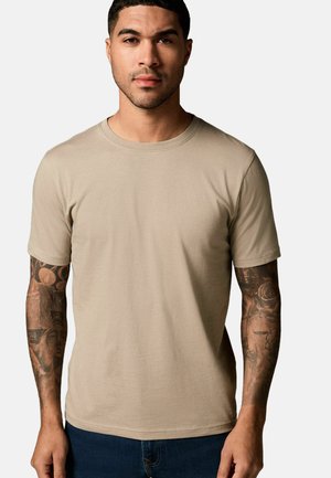 T-shirt in cotone beige con maniche corte e scollo rotondo. Texture liscia, design aderente e tatuaggi visibili sulle braccia.