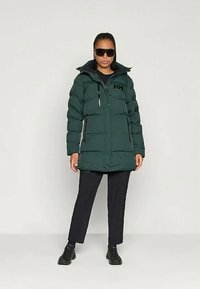 Πληρωμένο πράσινο παρκά μπουφάν με ζικ ζακ μπροστά, μαύρες λεπτομέρειες, ψηλό γιακά και πλάγιες τσέπες. Φορεμένο με μαύρο παντελόνι και γυαλιά ηλίου.