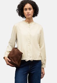 Blusa beige chiaro con colletto arricciato, bottoni frontali e orlo con coulisse. Abbinata a jeans blu e una pochette in suede marrone.