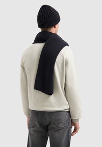 Zwarte gebreide beanie en bijpassende sjaal gedragen over een lichtbeige sweatshirt. Grijze jeans maken de outfit compleet en benadrukken zachte texturen en kleuren.