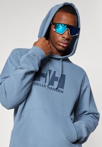 Felpa con cappuccio di colore azzurro realizzata in cotone, con una tasca frontale, caratterizzata da un grande logo scuro "HH" e il testo "HELLY HANSEN" sottostante. Modello che indossa occhiali da sole specchiati.