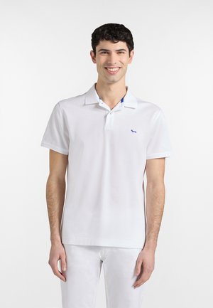 Giovane sorridente con capelli corti e ricci che indossa una polo bianca a maniche corte con un piccolo logo blu a forma di cane sul petto.