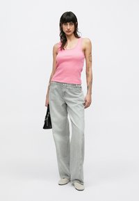 Camiseta de tirantes rosa con textura acanalada, vaqueros de pierna ancha en gris claro, bolso negro, zapatos claros. Varias tatuajes visibles en los brazos. Fondo sencillo.