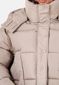Beige puffjacka med hög krage, med stora quiltade sektioner, tryckknappar och justerbara axelremmar. Slät, glänsande yta.