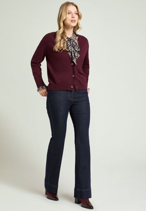 Kvinde iført en bordeaux cardigan, ternet bluse med sløjfe, mørkeblå jeans og bordeaux hæle, står mod en ensartet lys baggrund.