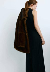 Manteau en fourrure marron à texture douce, drapé sur une robe noire sans manches. Le manteau présente une forme arrondie et est porté sur une épaule.