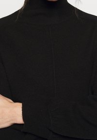 Svart polotröja med ribbat mönster, med långa ärmar och en åtsittande design. Sömdetalj som går vertikalt ner längs framsidan.