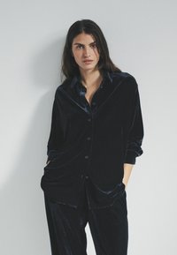 Schwarzes Samt-Hemd mit Knopfleiste, langen Ärmeln und lockerer Passform. Besitzt einen Kragen und Vordertaschen, kombiniert mit passenden Satin-Hosen.