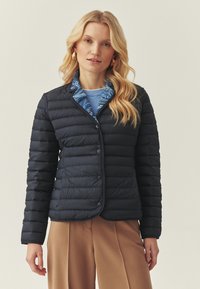 Femme aux longs cheveux blonds portant une veste matelassée bleu marine à boutons sur une chemise bleue et un pantalon large couleur sable, debout devant un fond uni.